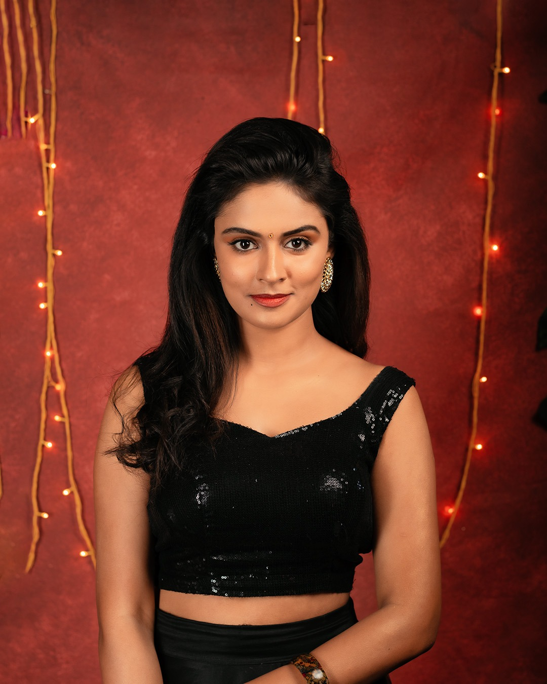 Woman wearing a black sequin crop top with voluminous side-swept hair and elegant soft glam makeup against a warm red studio background