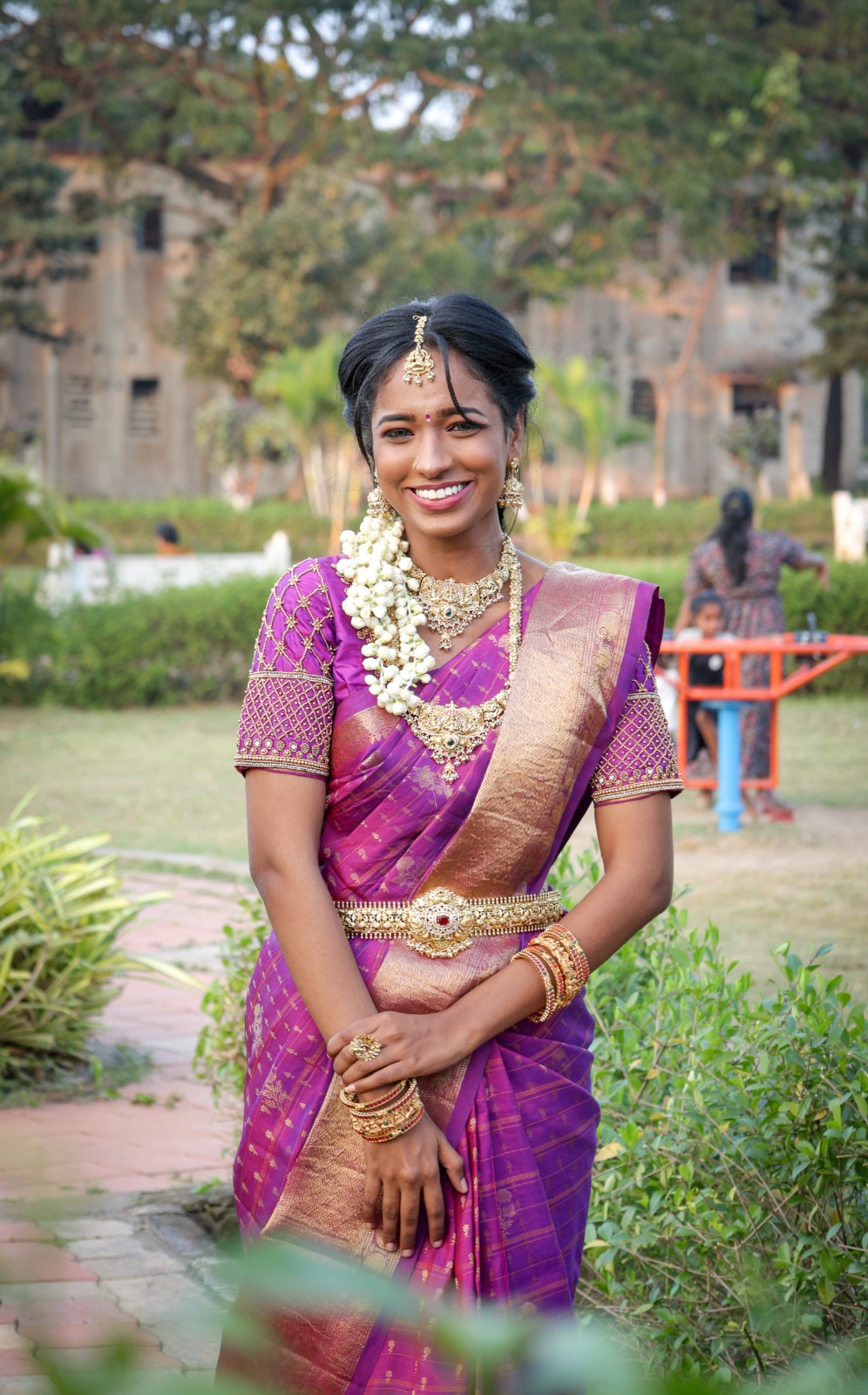 South Indian bride wearing a purple silk saree with a gold border, traditional gold temple jewellery, jasmine flowers, and elegant bridal makeup in an outdoor setting