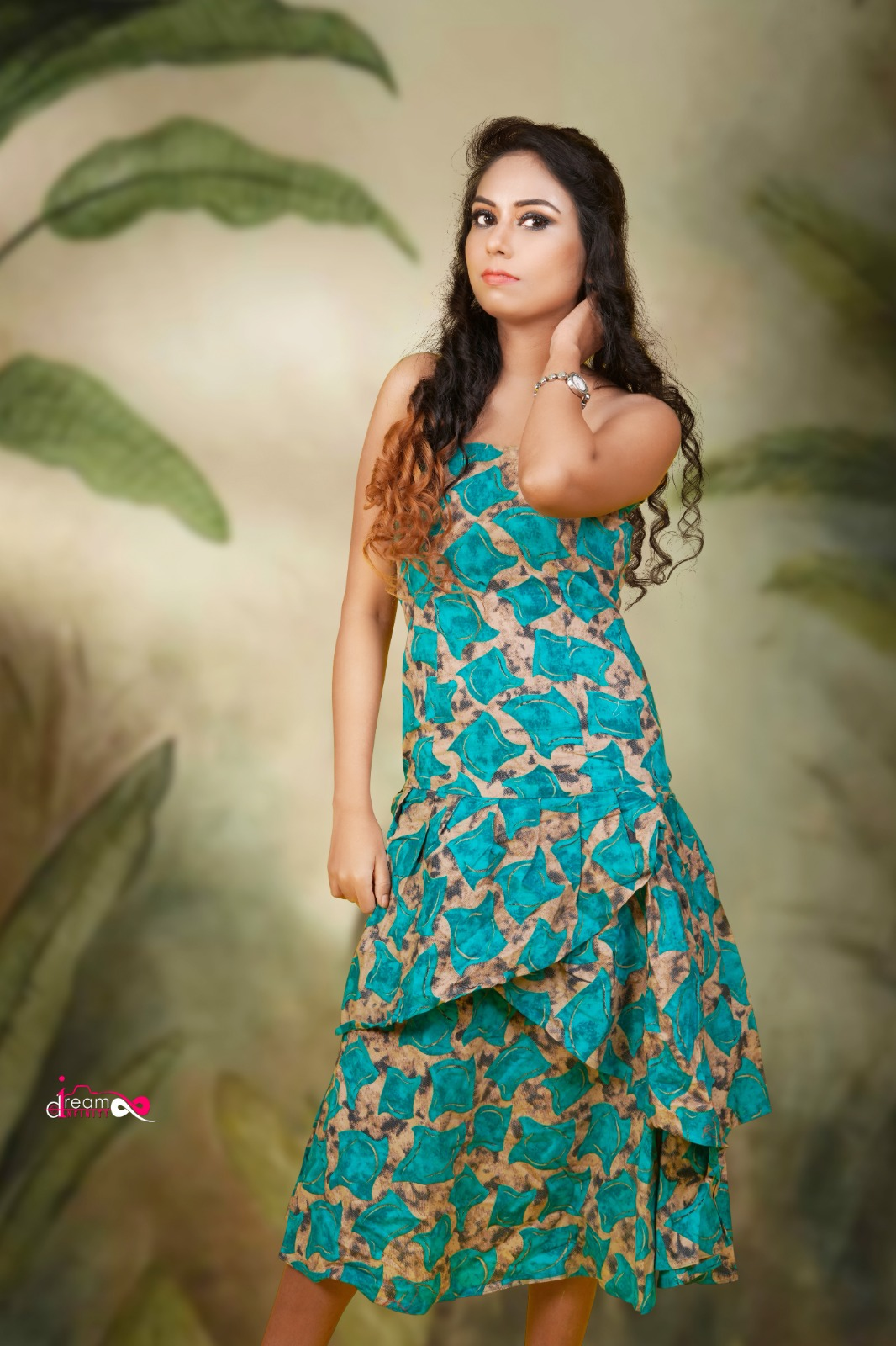 Woman wearing a teal blue patterned layered gown with long wavy hair and soft glam makeup against a natural studio background